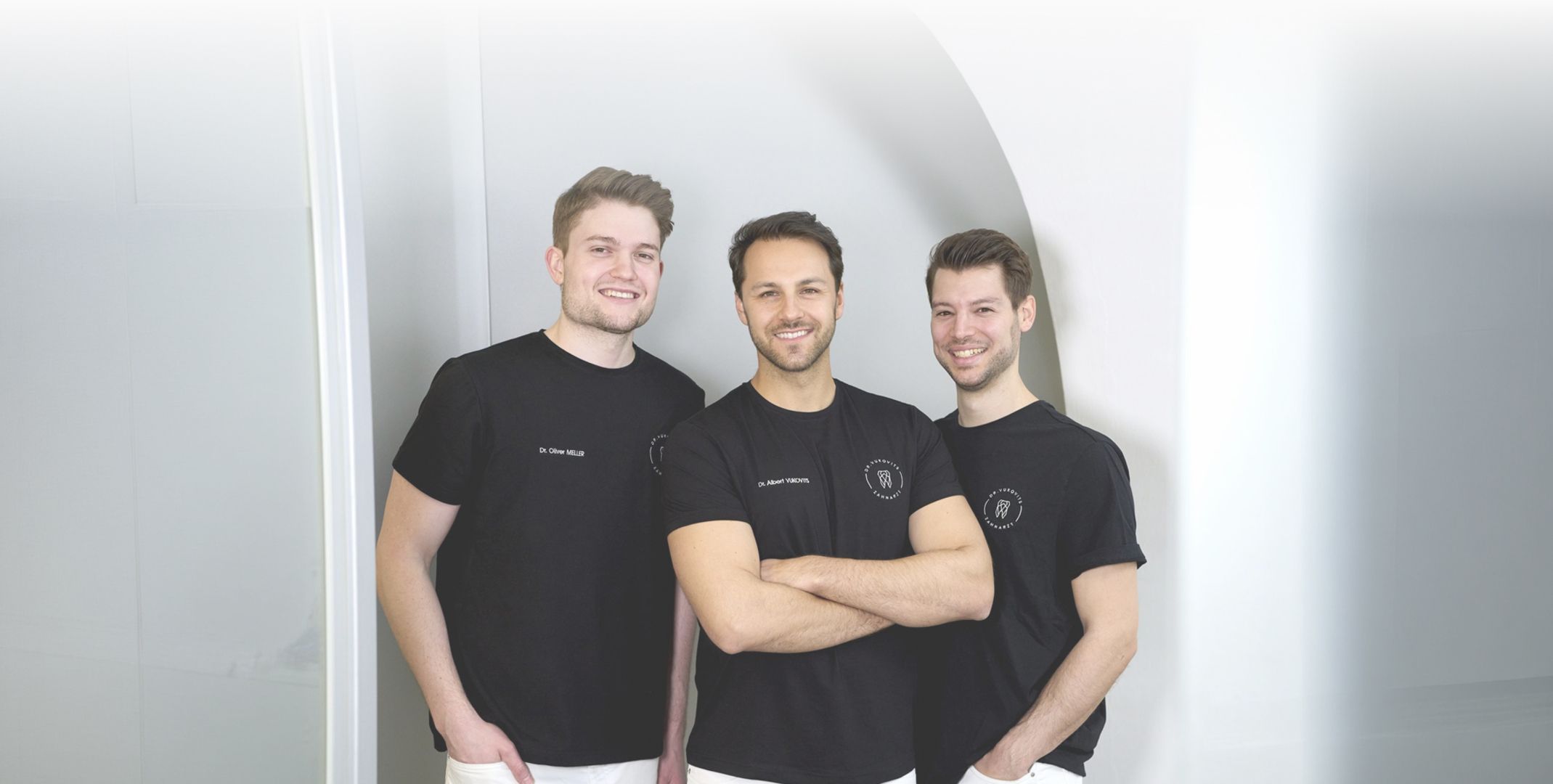 Drei Männer in schwarzen T-Shirts mit weißer Schrift stehen vor einer hellen Wand, einer mit verschränkten Armen in der Bildmitte